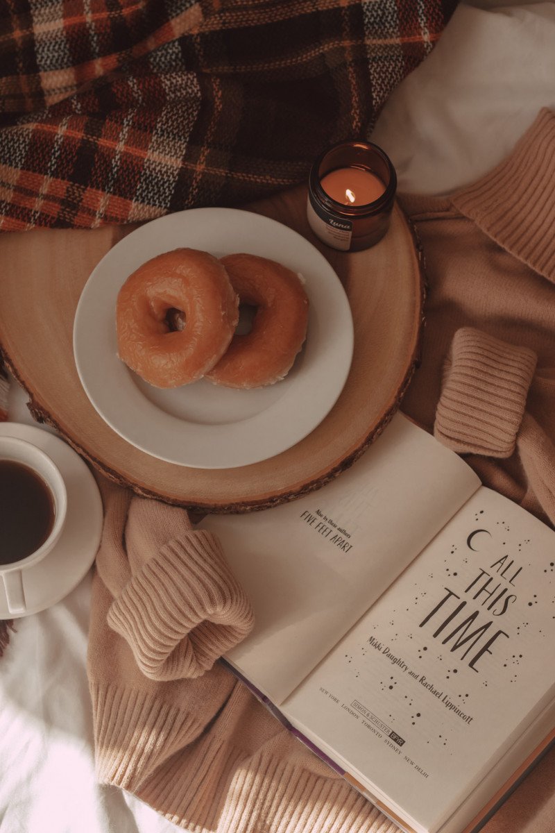 coffee mug and donuts laid on top of a taupe sweater with all this time book open to title page