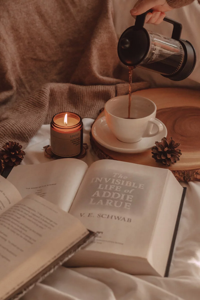 coffee being poured into a mug next to a lit candle with two open books int he foreground