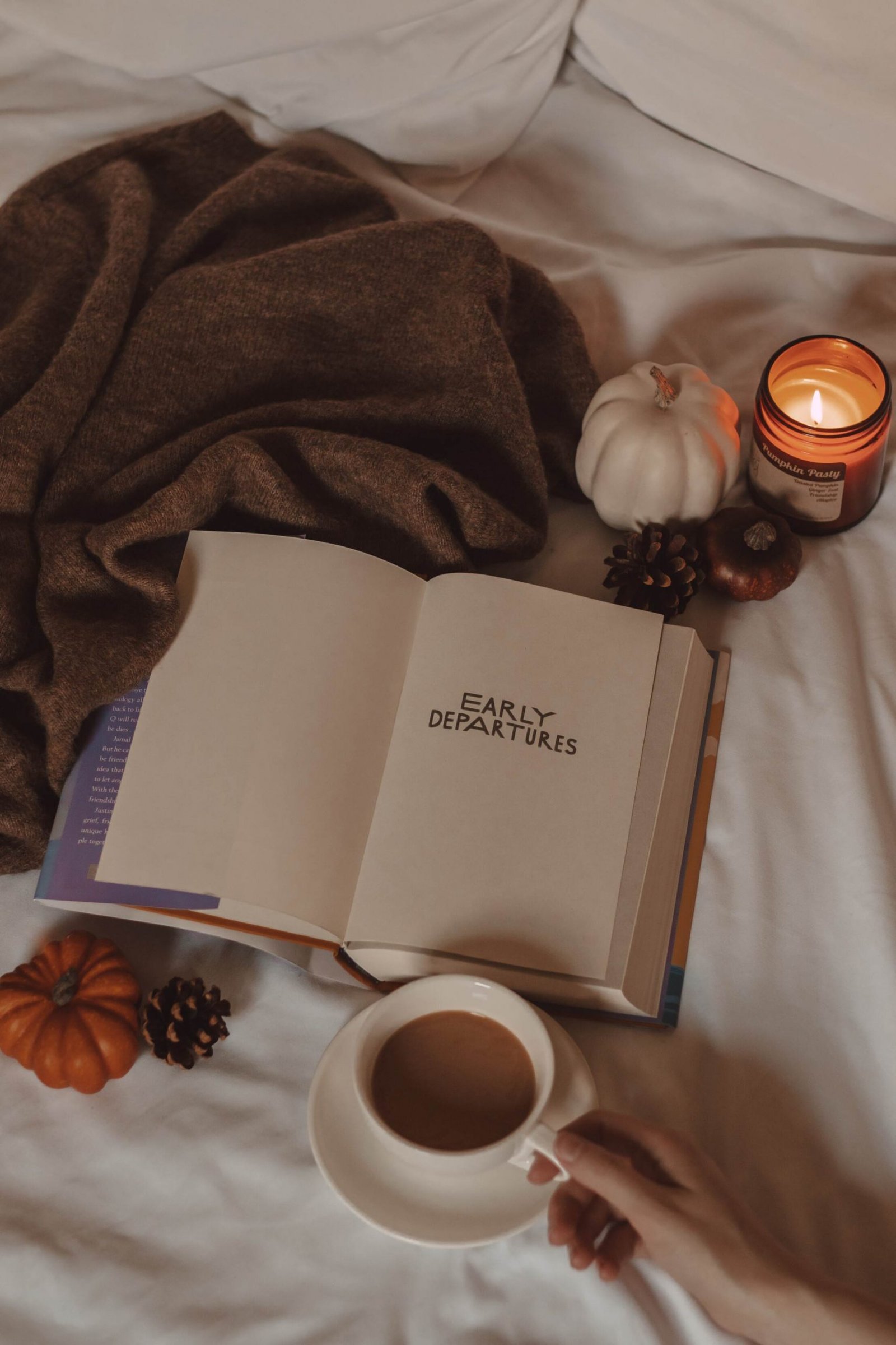 early departures book lying open with a mug of coffee, a sweater, mini pumpkins, and a lit candle