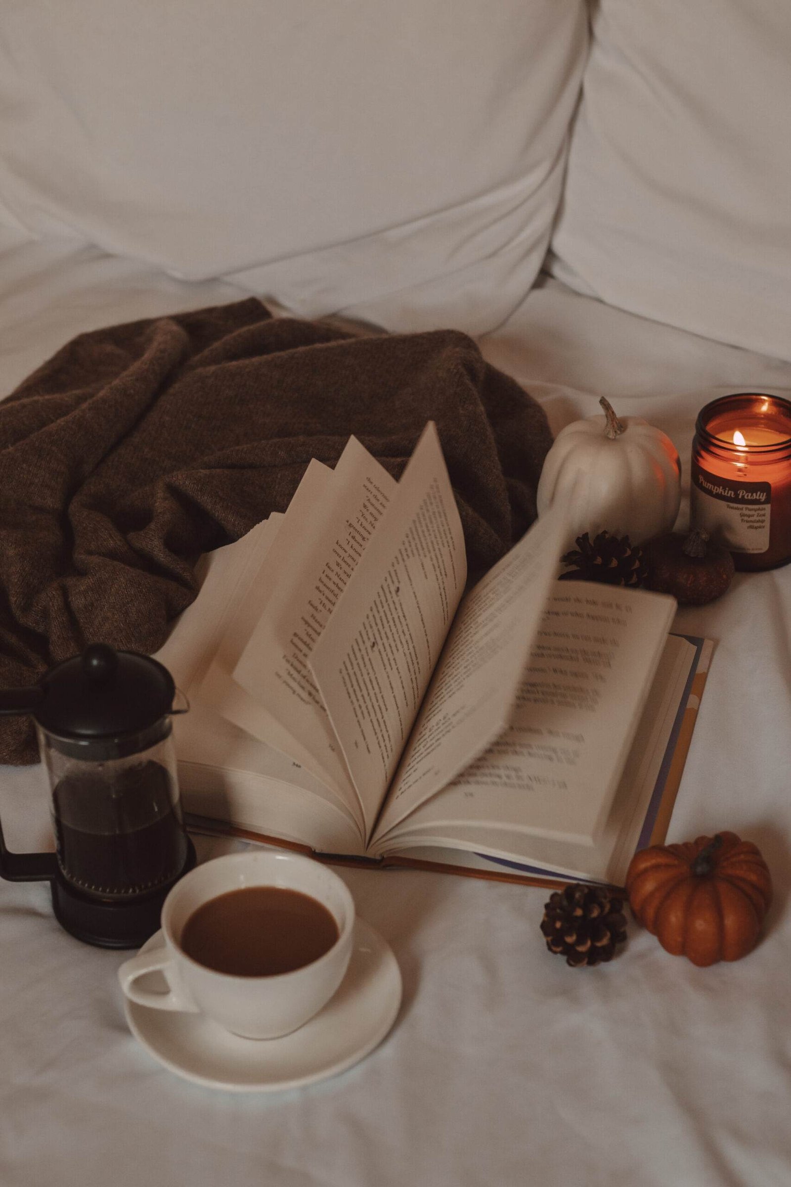 pages flipping in a book next to a french press and mug of coffee, mini pumpkins, and a candle