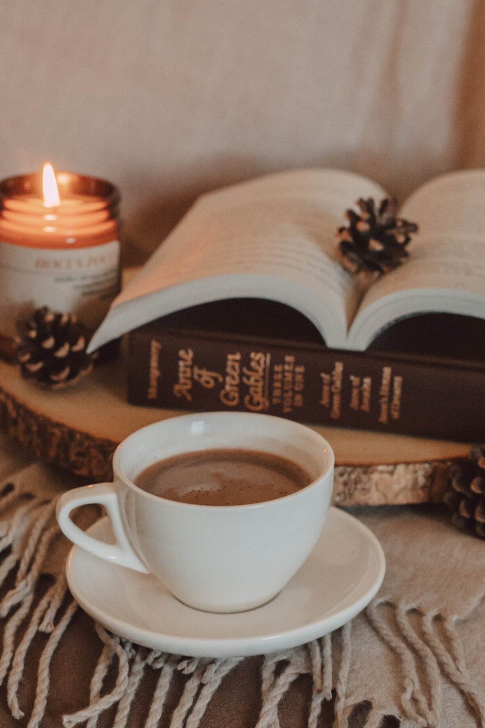 mug of mocha in front of vintage copy of anne of green gables book with a burning candle and pinecones