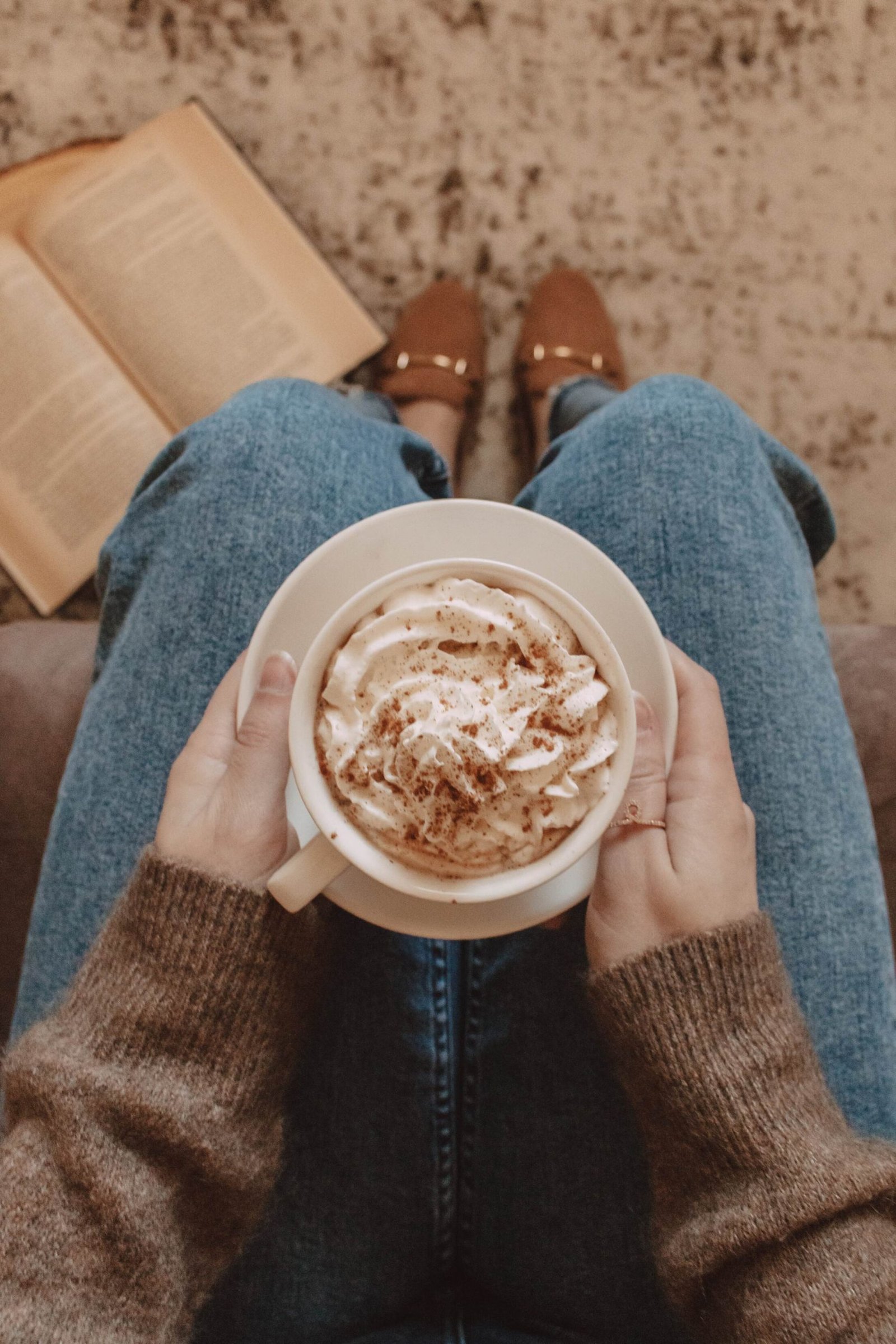 hands holding a mug filled with whipped cream and cinnamon on top of lap with book laying on the floor