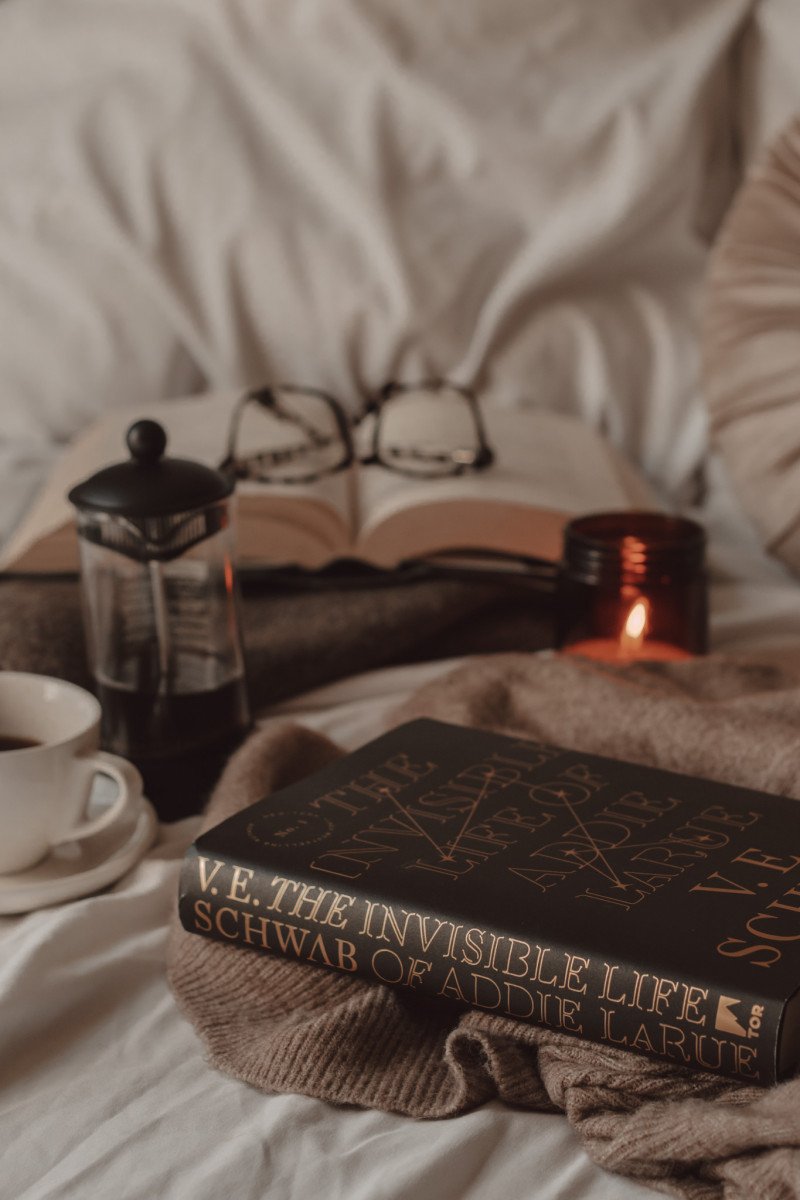 the spine of the invisible life of addie larue by v.e. schwab next to a french press and mug of coffee with candle and open book with glasses on top