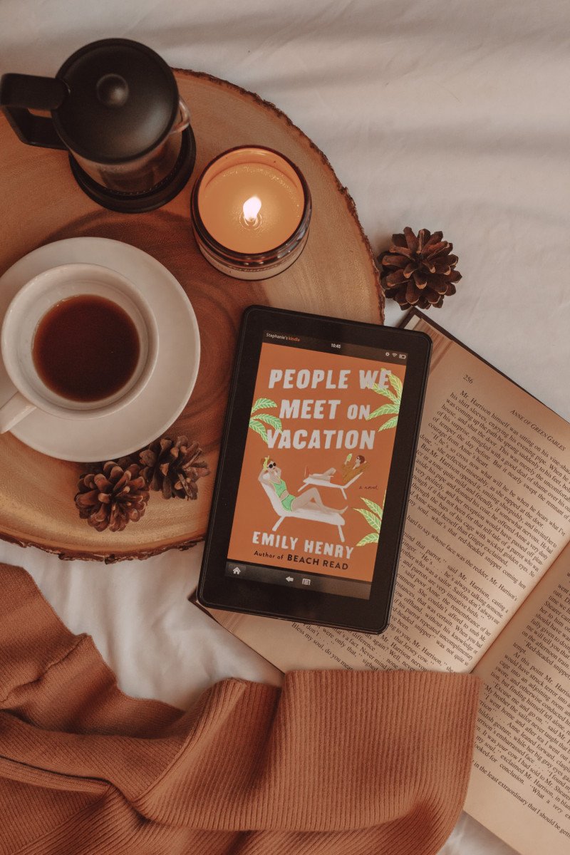 from above, you can see an e-reader with the book cover for "people we meet on vacation" laying on an open book. next to it are a mug of coffee, a french press, and a lit candle.