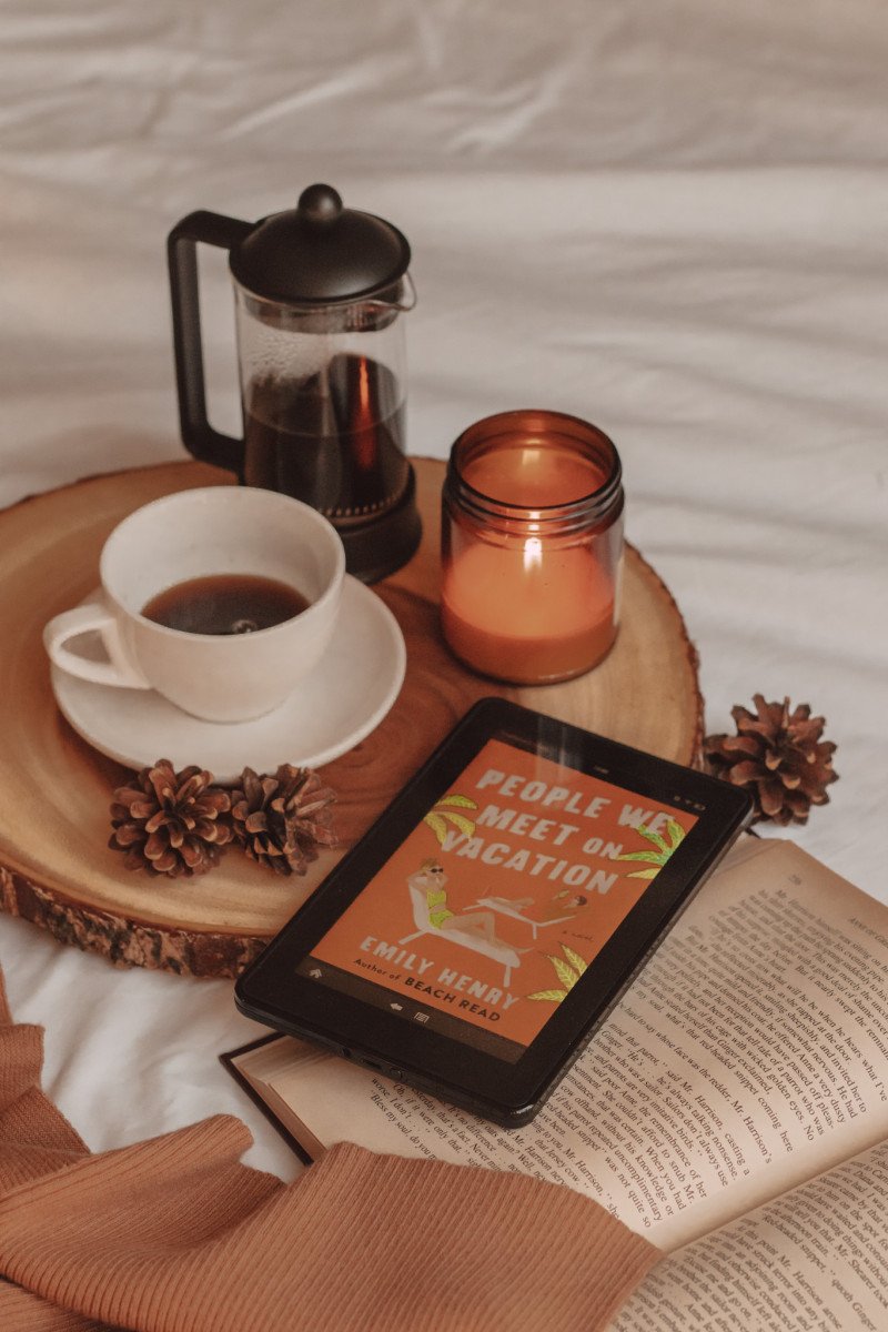 a kindle sits on top of an open book with "people we meet on vacation" shown on the screen. a lit candle, mug of coffee, and french press sit beside it.