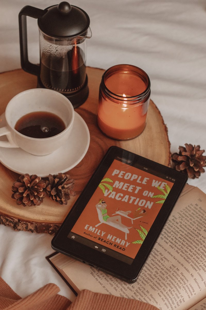 kindle with the cover of "people we meet on vacation" shown on the screen. a mug of coffee, a french press, and a lit candle sit beside it