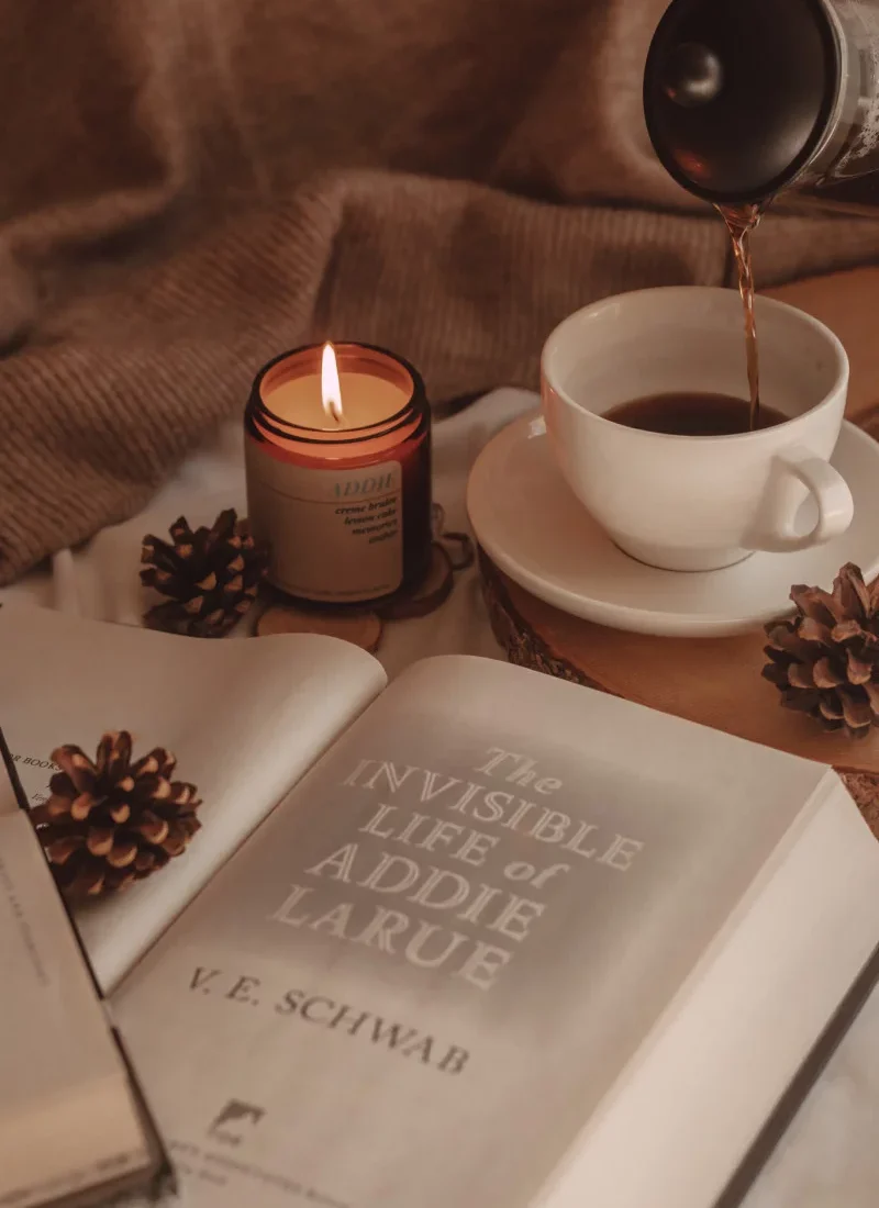 a close-up of the title page of the invisible life of addie larue with a lit candle and coffee being poured into a mug in the background