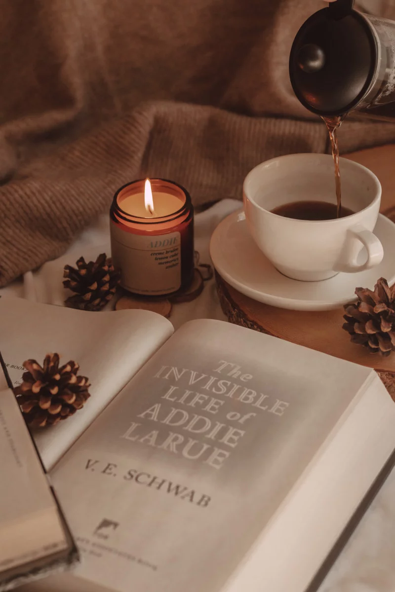 a close-up of the title page of the invisible life of addie larue with a lit candle and coffee being poured into a mug in the background