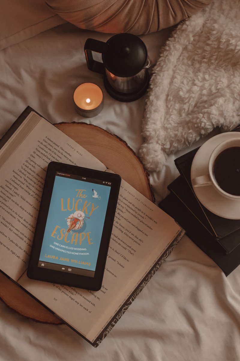The Lucky Escape book cover shows on a Kindle screen as it lays on an open book next to a mug of coffee on top of a stack of books and a lit candle next to a french press