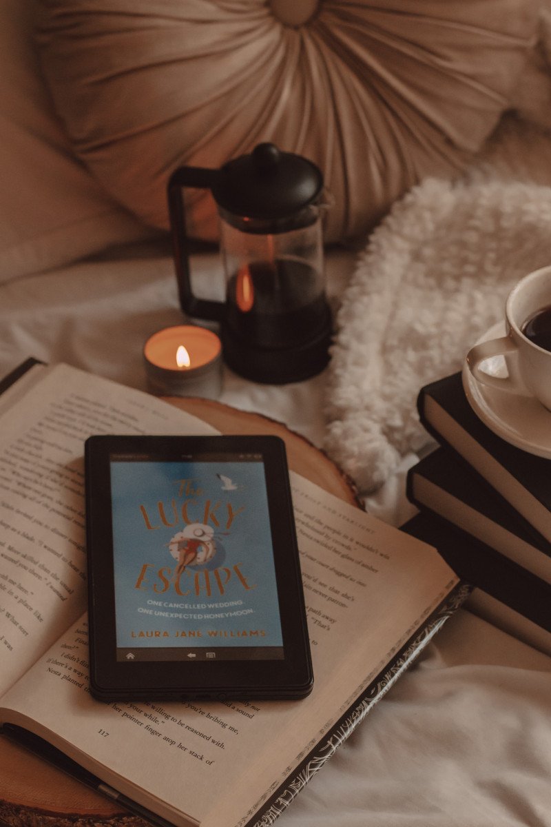 kindle sits face up on top of an open book with The Lucky Escape book cover on the screen. a small tin candle is lit next to a french press in the background and a mug of black coffee sits on three black books next to it all.