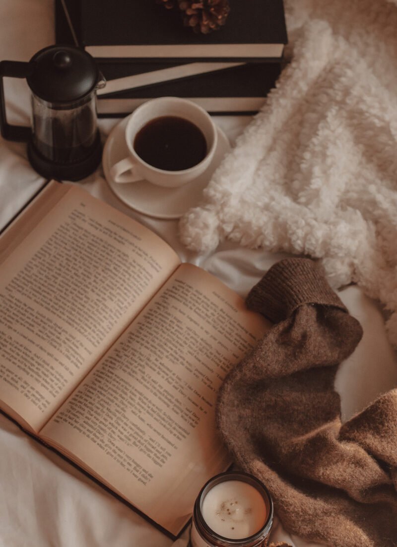 vintage book lies open with a mug of black coffee and french press next to a stack of black books and the arm of a sweater