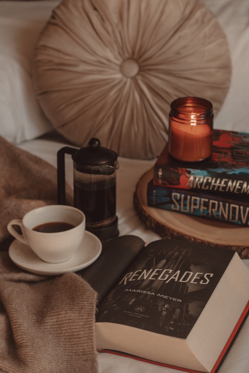 Renegades book open to title page next to a mug of coffee and French press. Archenemies and Supernova books stacked in the background with a lit candle on top