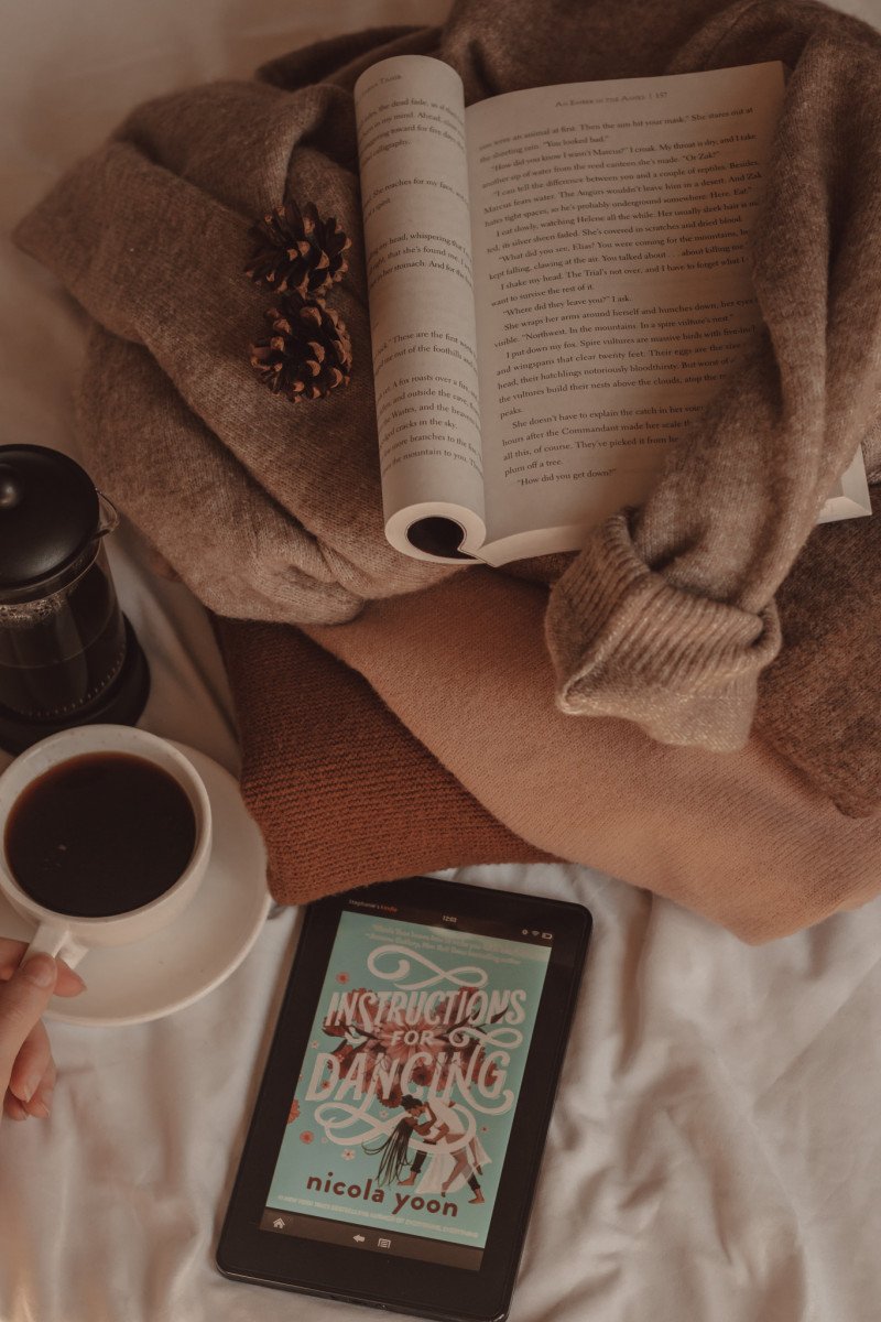 Instructions for Dancing book cover visible on e-reader with a cup of coffee next to a french press. A book is folded open on top of a pile of sweaters.