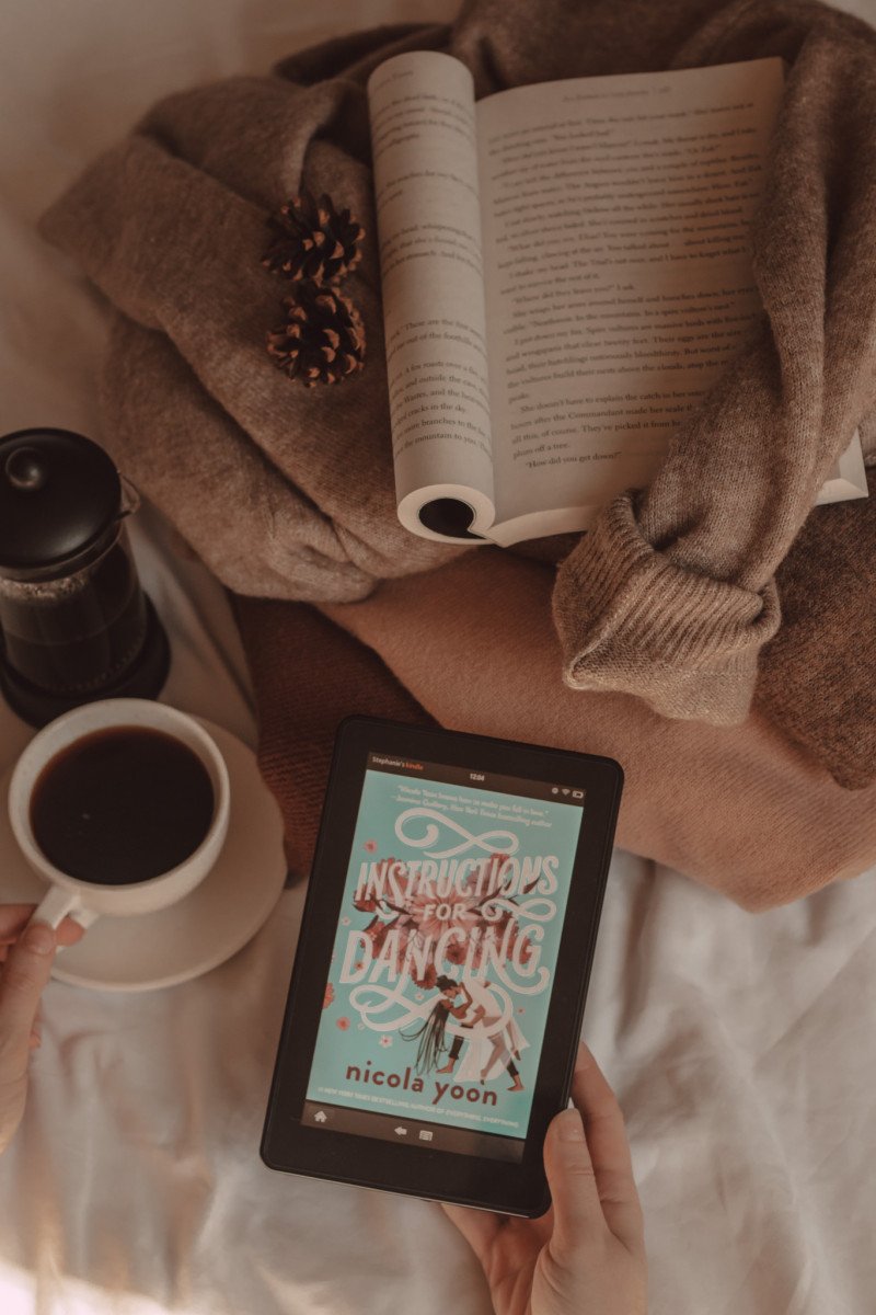 An e-reader shows the cover of Instructions for Dancing by Nicola Yoon while a book lays on top of a stack of sweaters and a hand holds a mug of coffee next to a French press.