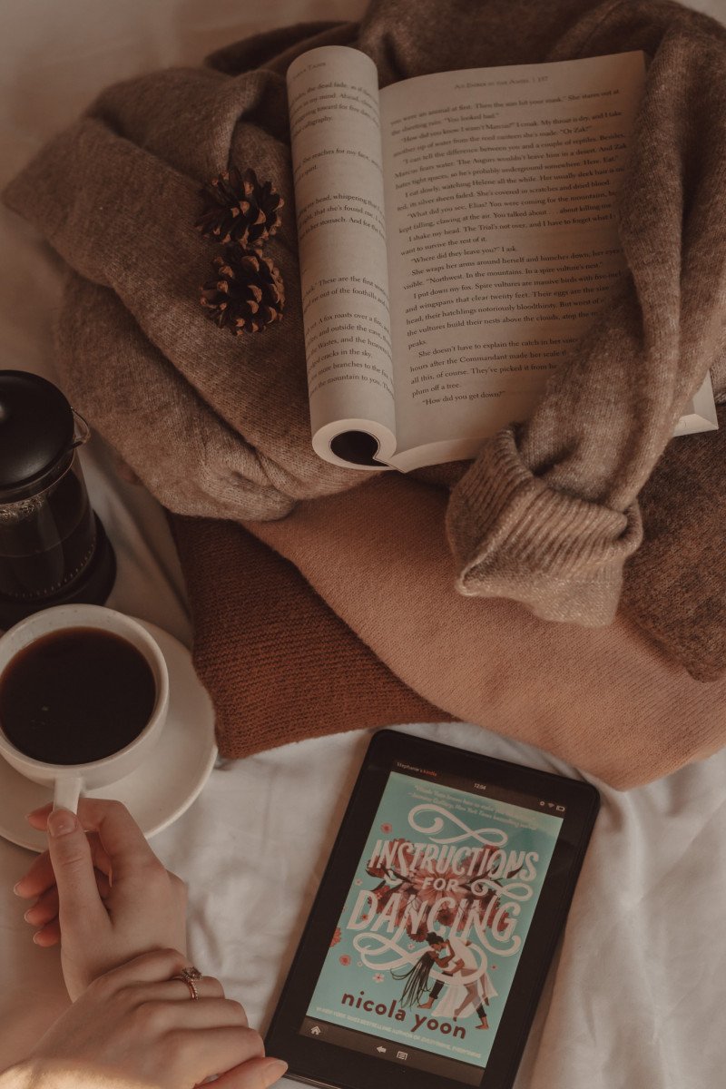 A hand holds a mug of black coffee next to a French press and a Kindle with the cover of Instructions for Dancing visible on the screen. Next to them, a stack of sweaters has a paperback book folded open on top of it.