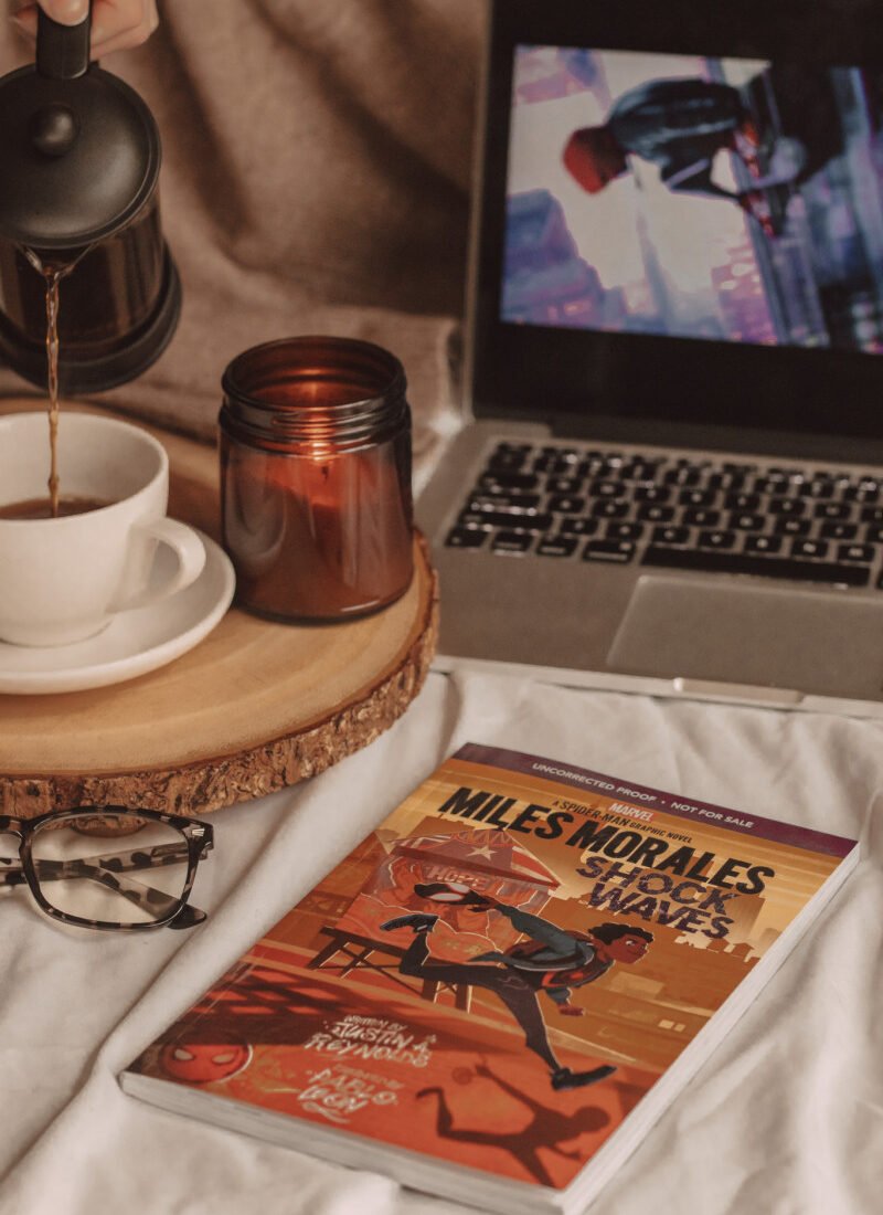 graphic novel of miles morales sits cover-up in the foreground next to a pair of glasses, a coffee mug, and a candle. laptop open to reveal screen in the background