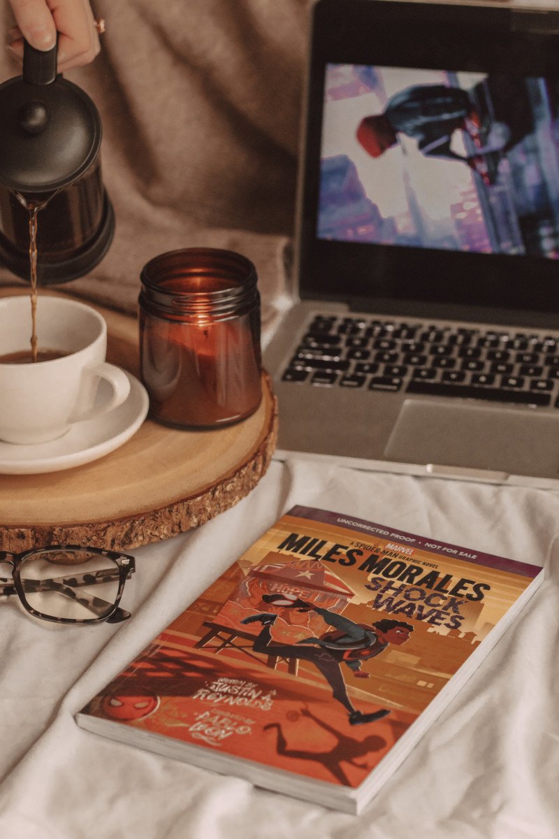 graphic novel of miles morales sits cover-up in the foreground next to a pair of glasses, a coffee mug, and a candle. laptop open to reveal screen in the background