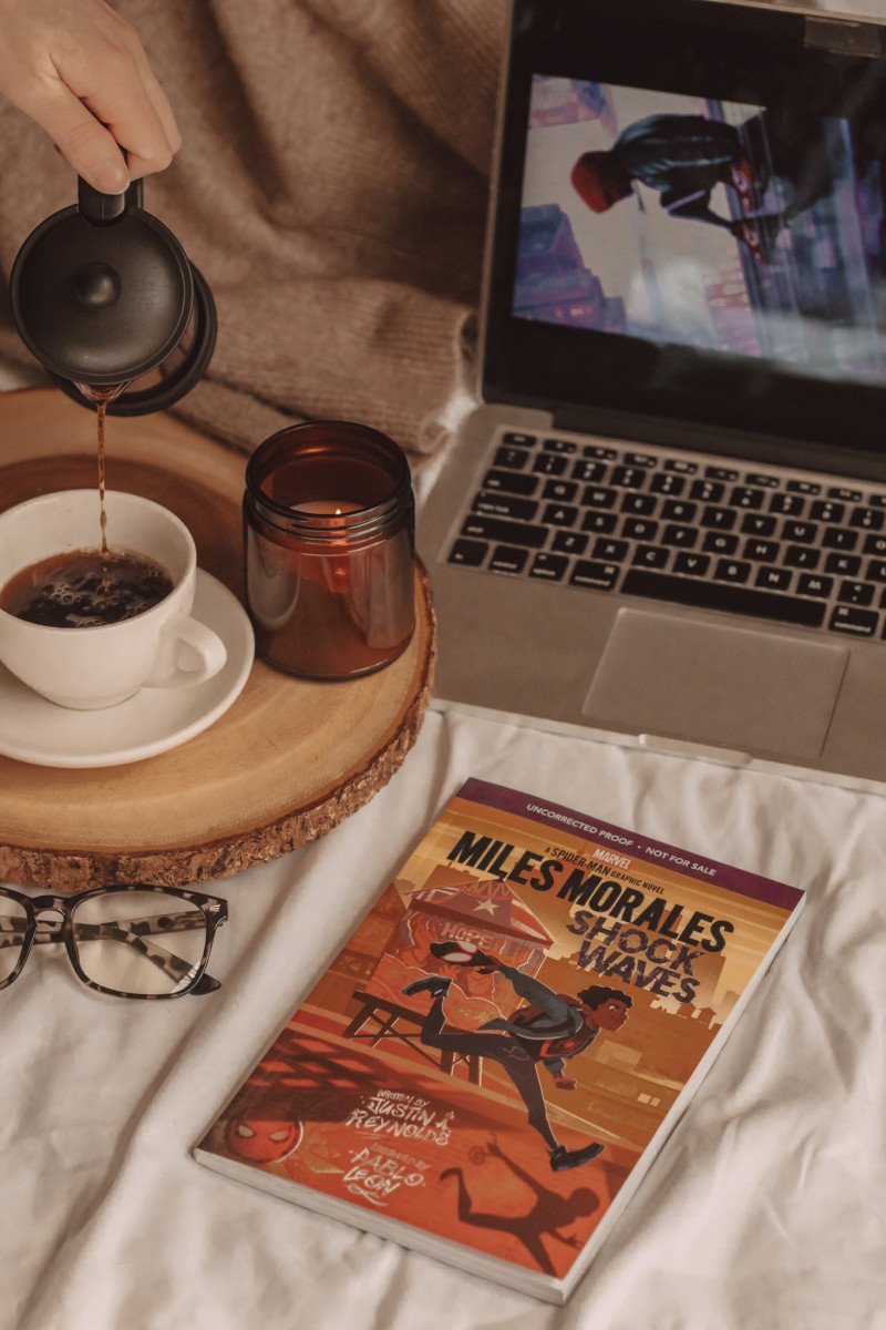 miles morales graphic novel sits next to a pair of glasses and a laptop while a hand pours coffee into a mug next to a candle