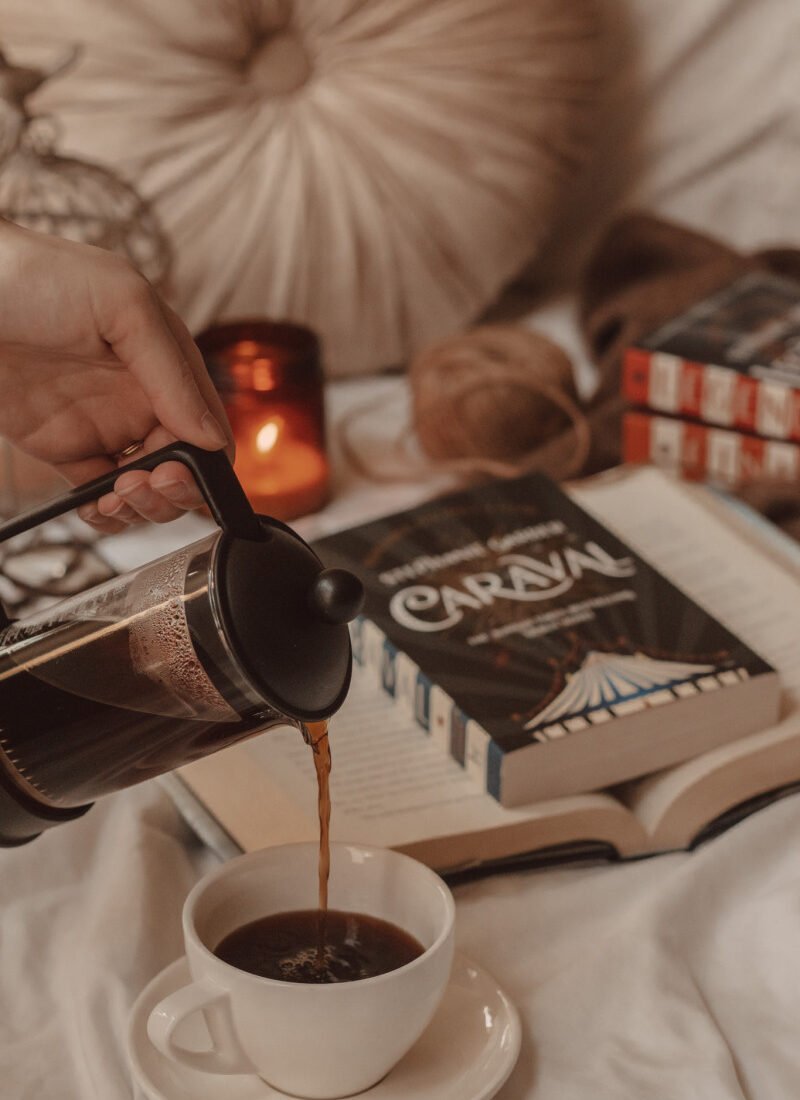 black coffee poured from a french press into a mug in the foreground with Caraval in the background laying on an open book with a lit candle burning next to it