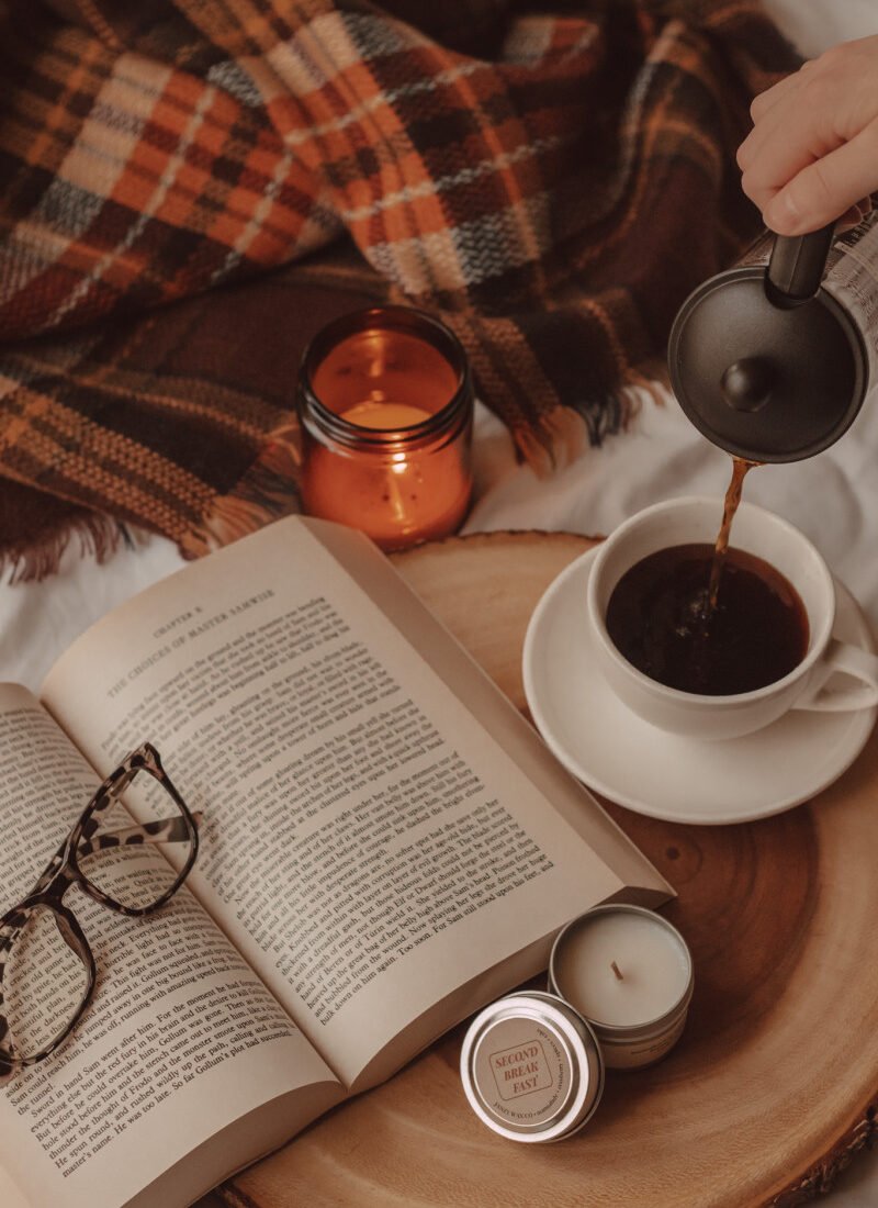a hand pours black coffee into a mug next to an open book with glasses laying on top. a lit candle in a jar is in the background with a plaid scarf and a small, unlit candle tin is in the front on a wooden cake tray.