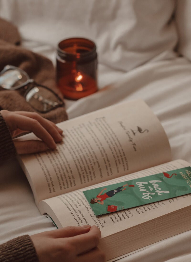 bookmark lays on an open book while a hand holds the book open and a candle burns in the background