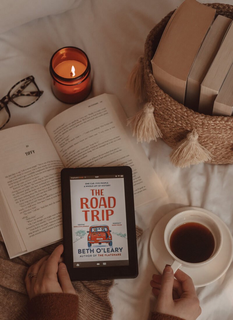hands grip a mug of coffee and e-reader with The Road Trip book cover on it. beneath lies an open book and next to it are a basket of books and a lit candle