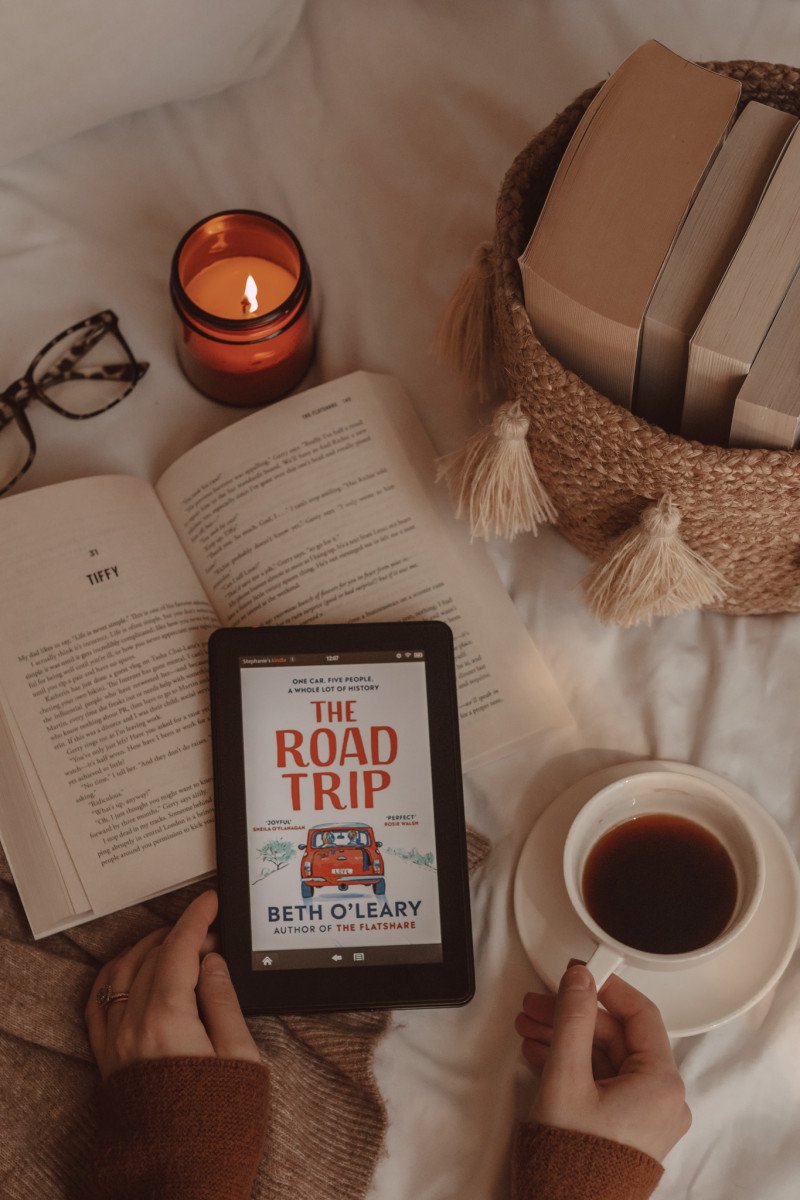 hands grip a mug of coffee and e-reader with The Road Trip book cover on it. beneath lies an open book and next to it are a basket of books and a lit candle