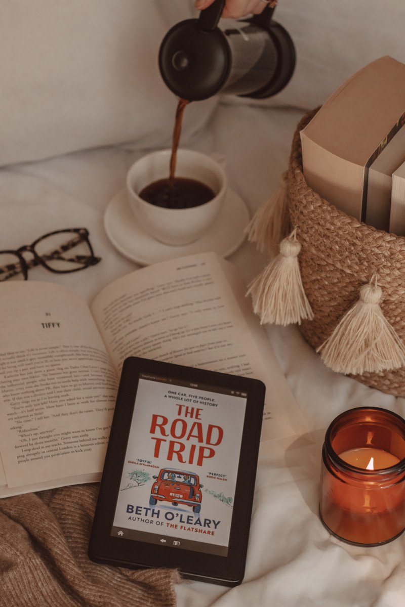 an e-reader is in the foreground with The Road Trip book cover on the screen. next to it is a lit candle and behind it is a basket of books and a hand pouring black coffee into a mug