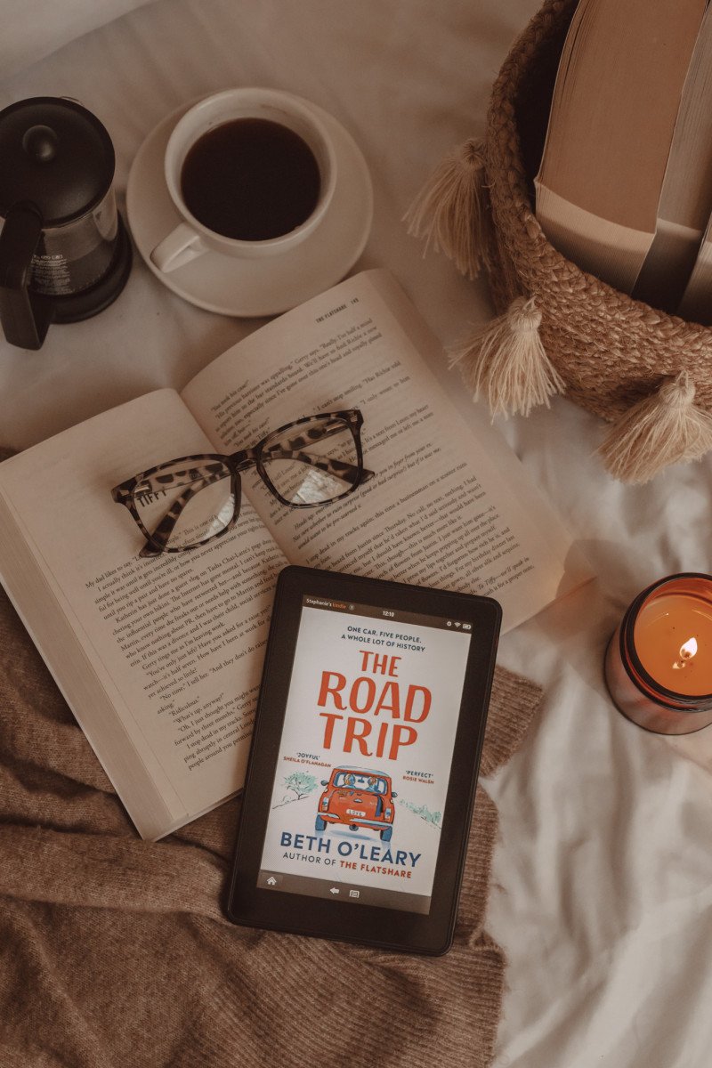 from above you see a kindle with The Road Trip book cover on it. it lies on an open book with a pair of glasses. next to it is a lit candle, a mug of black coffee, and a basket of books