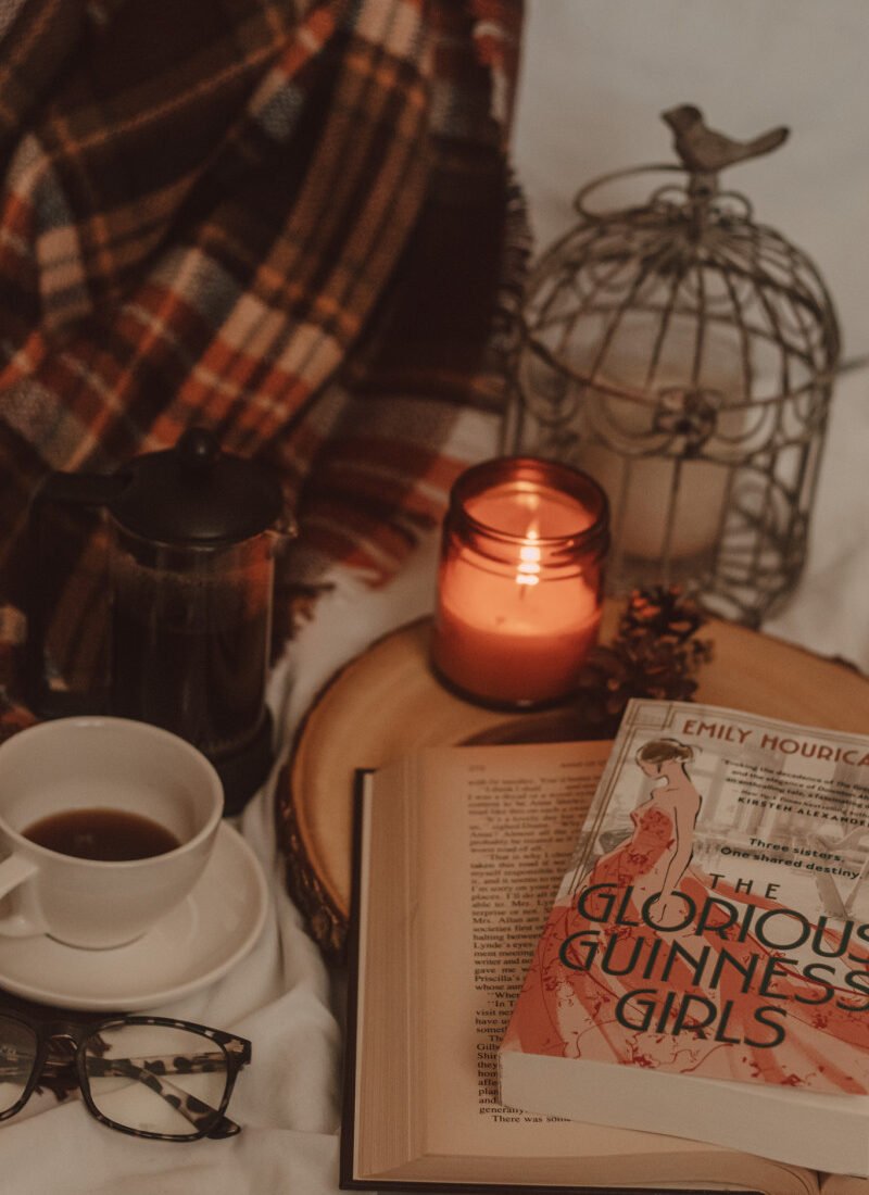 The Glorious Guinness Girls book sits atop an open book on a wooden cake tray next to a lit candle and mug of coffee