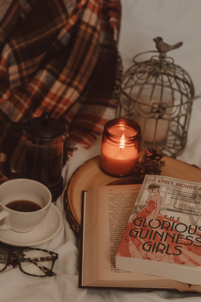 The Glorious Guinness Girls book sits atop an open book on a wooden cake tray next to a lit candle and mug of coffee