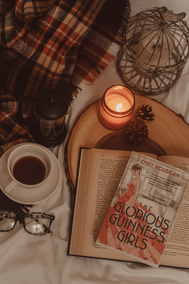 from above you see The Glorious Guinness Girls book on top of an open book on a wood tray next to a mug of coffee and lit candle