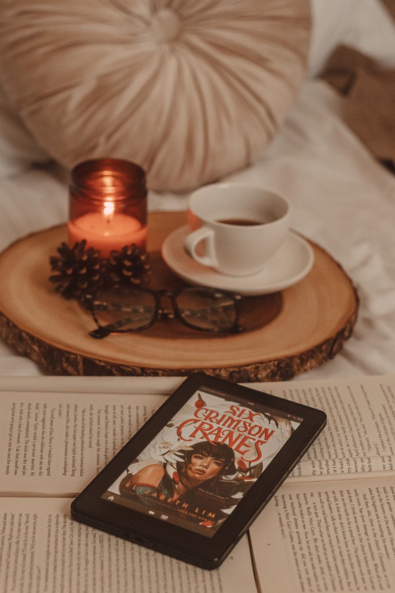 kindle with six crimson cranes book cover visible laying on top of several open books with a wooden board in the background, holding a mug and a lit candle