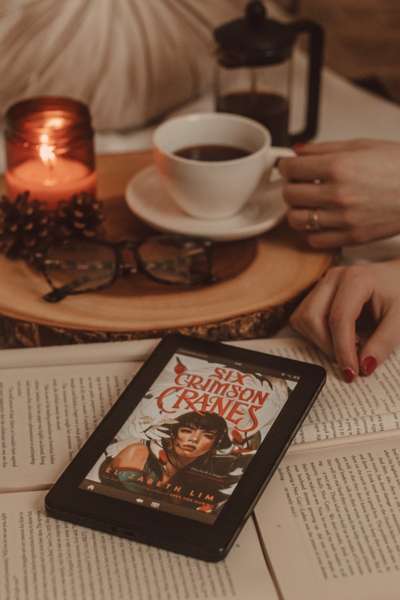 hands hold a mug of coffee next to a lit candle in the background while an ereader sits on top of open books with six crimson cranes cover visible on the screen.