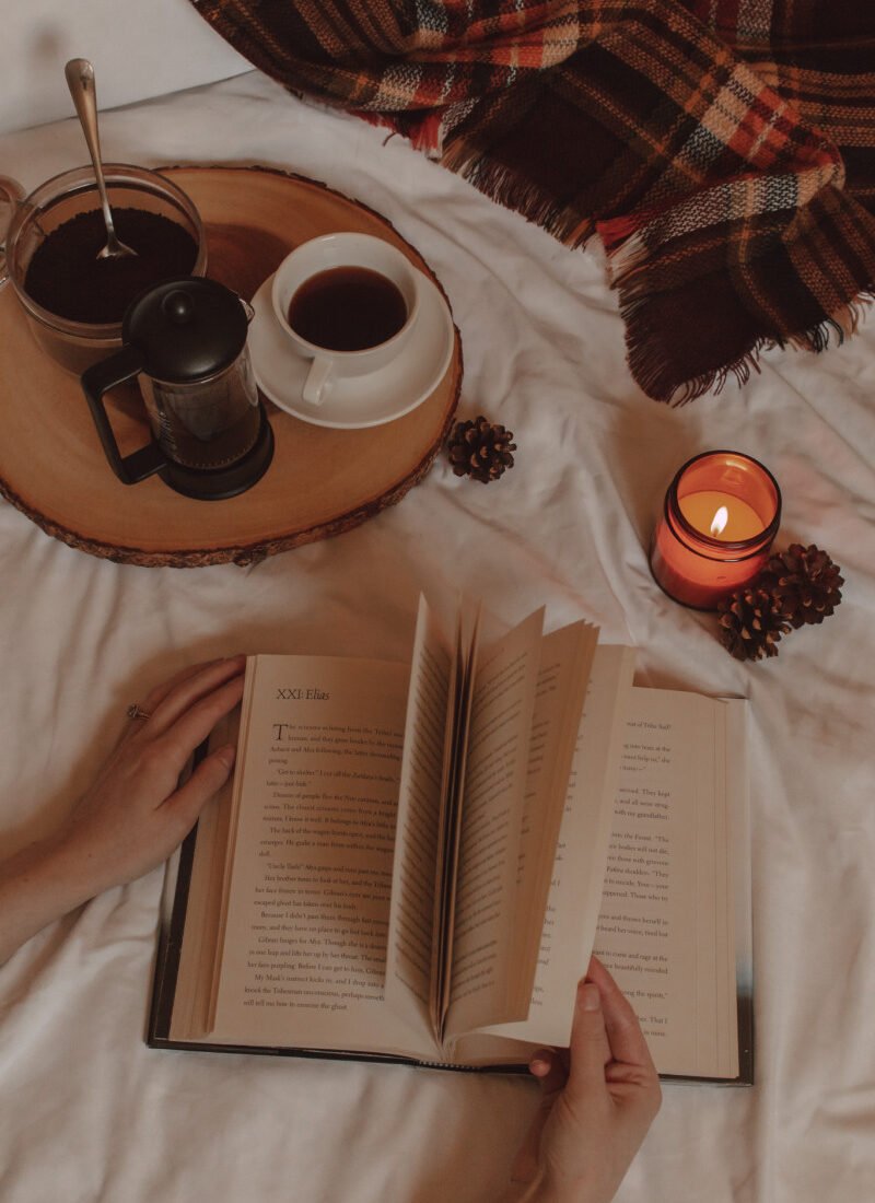 hands flip the pages of a book next to a burning candle and a mug of coffee